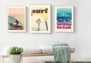 Three different surf posters and surfing art prints in wooden frames hanging on the wall above a white cushioned bench 